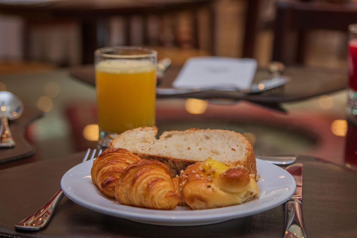Prato de café da manhã com croissant, pão e pastel, suco de laranja, sobre uma mesa de vidro.