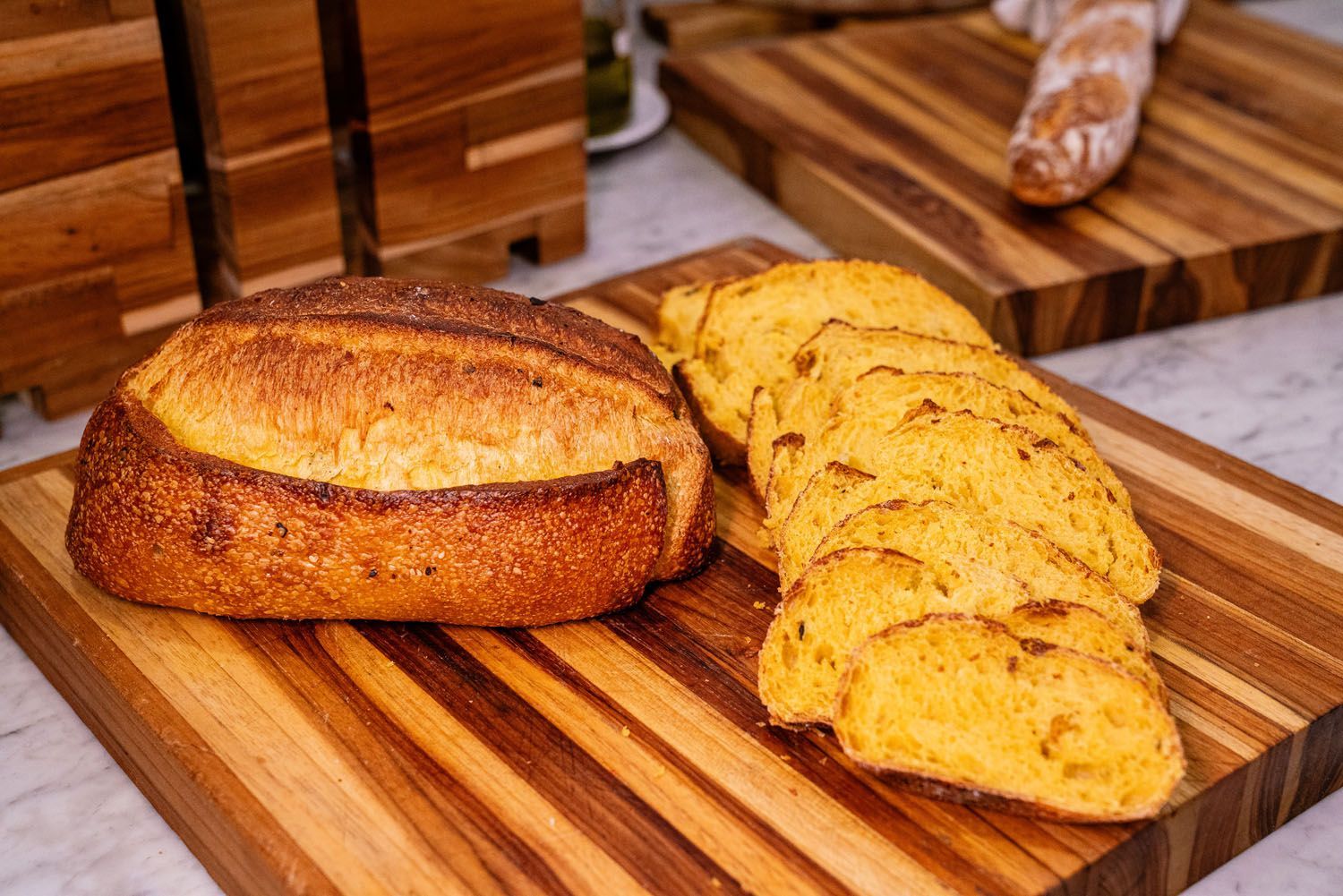 Pão de forma e fatias de pão sobre uma tábua de corte de madeira; mais pão ao fundo.