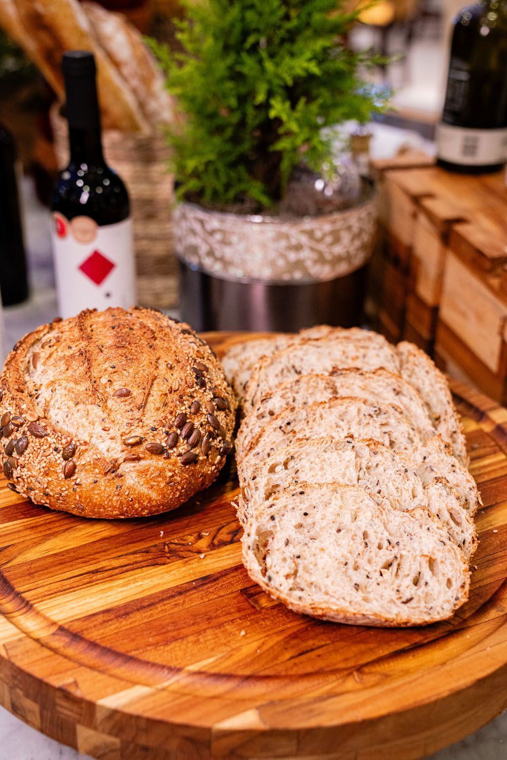 Pão de forma e fatias de pão sobre uma tábua de madeira, com garrafas de vinho e uma planta ao fundo.