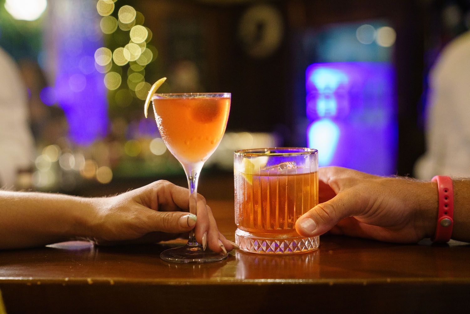 Duas mãos segurando coquetéis em um bar; uma em uma taça com haste e guarnição, a outra em um copo baixo.