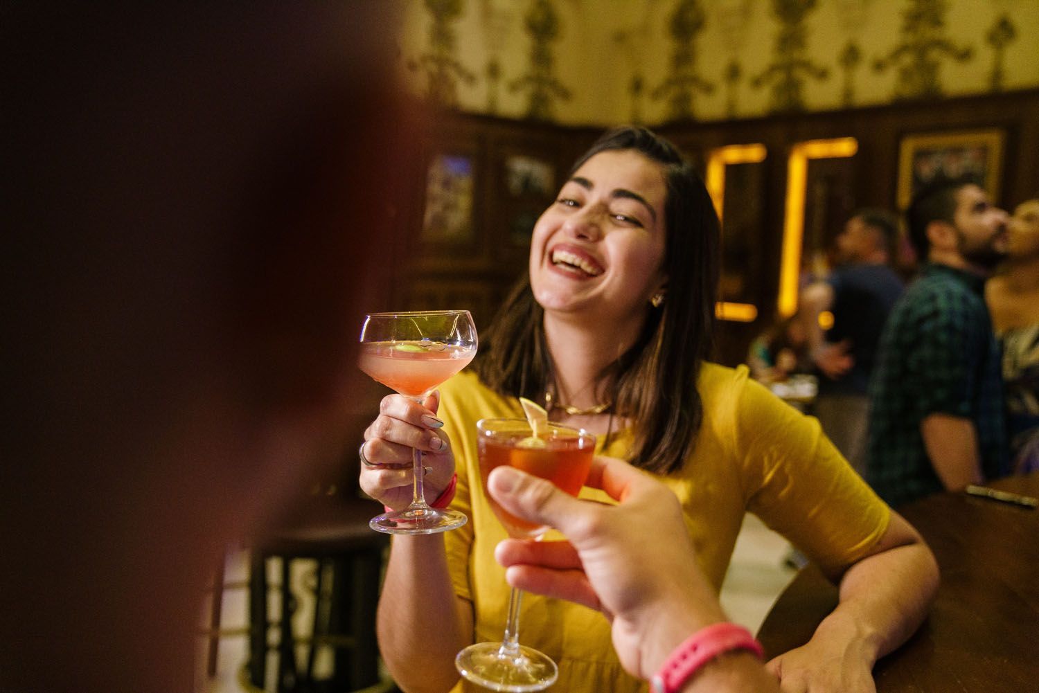 Mulher rindo, brindando com alguém em um bar. Ela veste amarelo; o cenário é de madeira escura.