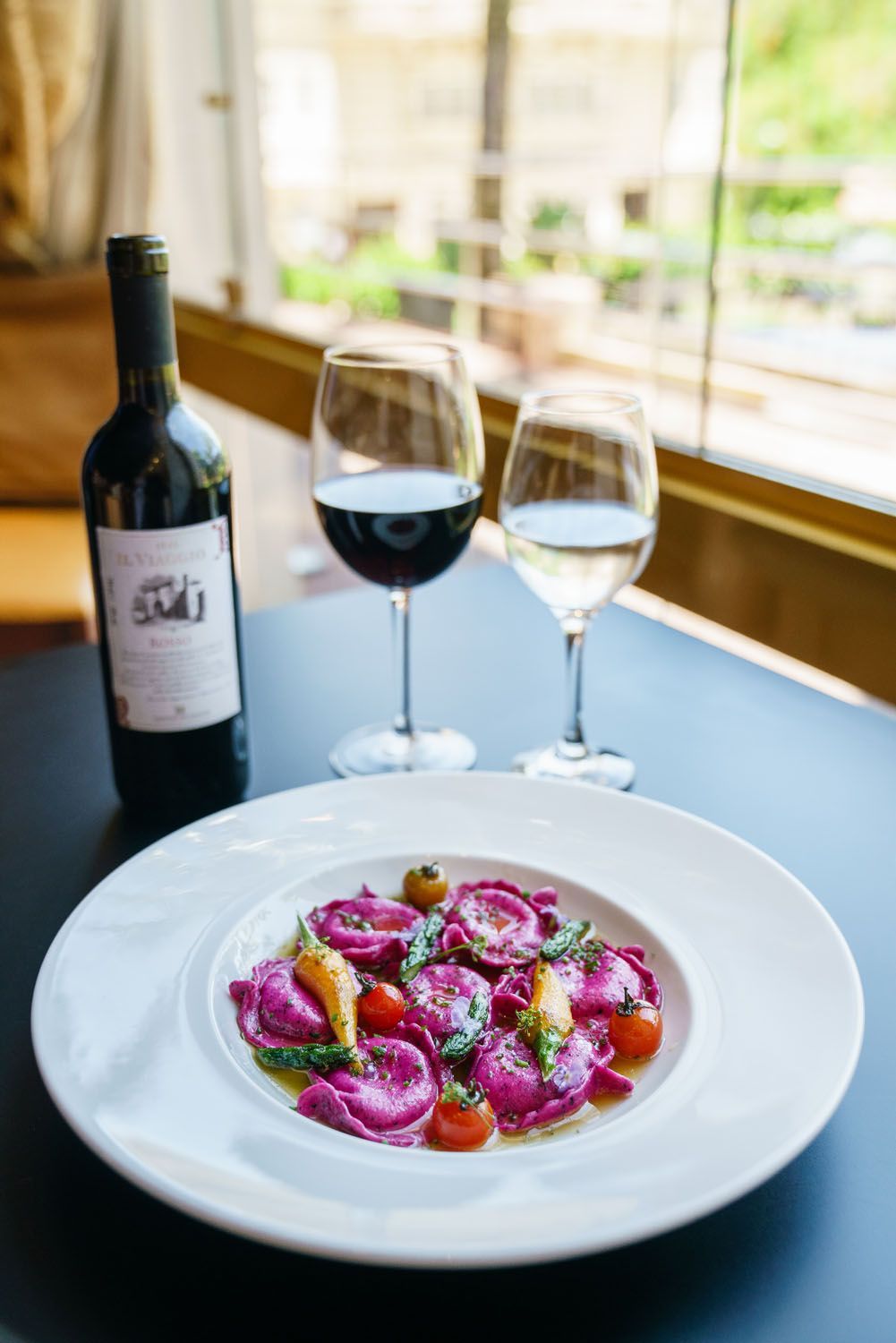 Prato de ravióli roxo com taças de vinho e uma garrafa de vinho sobre uma mesa perto de uma janela.