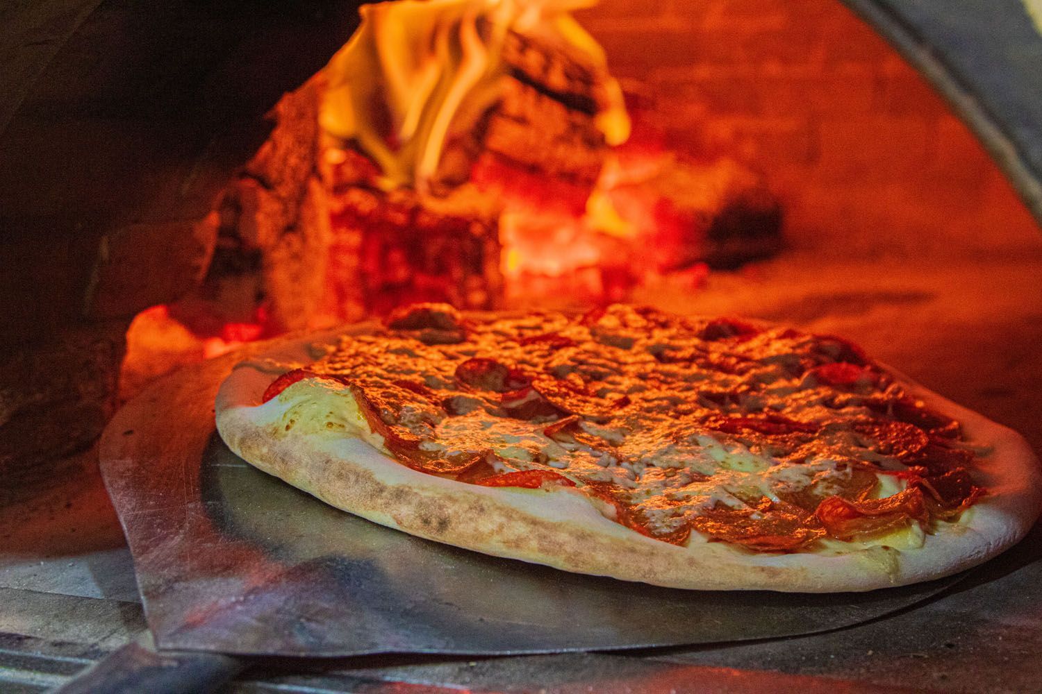 Pizza em forno a lenha, com chamas visíveis ao fundo, sendo assada em uma pá de madeira.