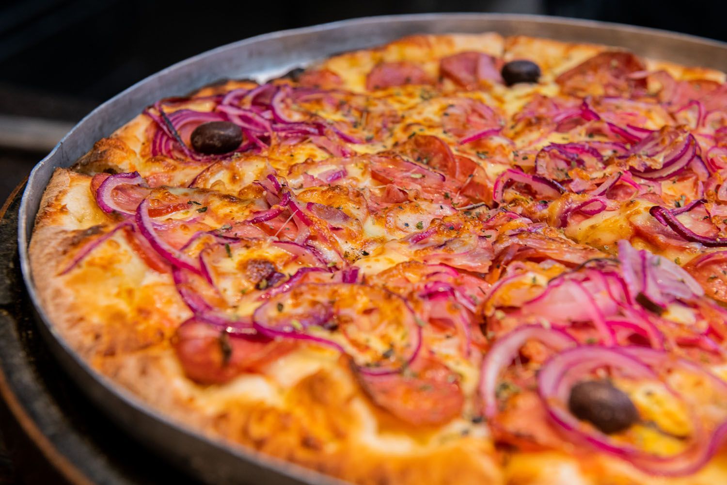 Pizza com cebola roxa, linguiça e azeitonas em uma assadeira de metal.