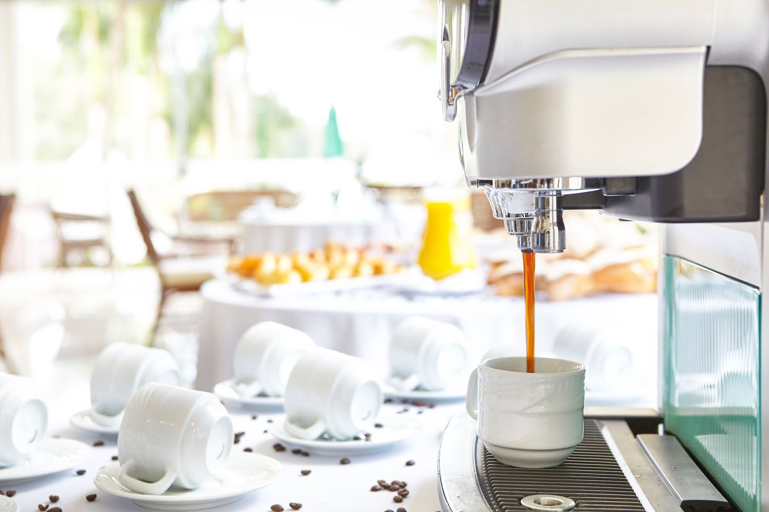 Máquina de café servindo café em uma xícara branca, em um ambiente de bufê de café da manhã.