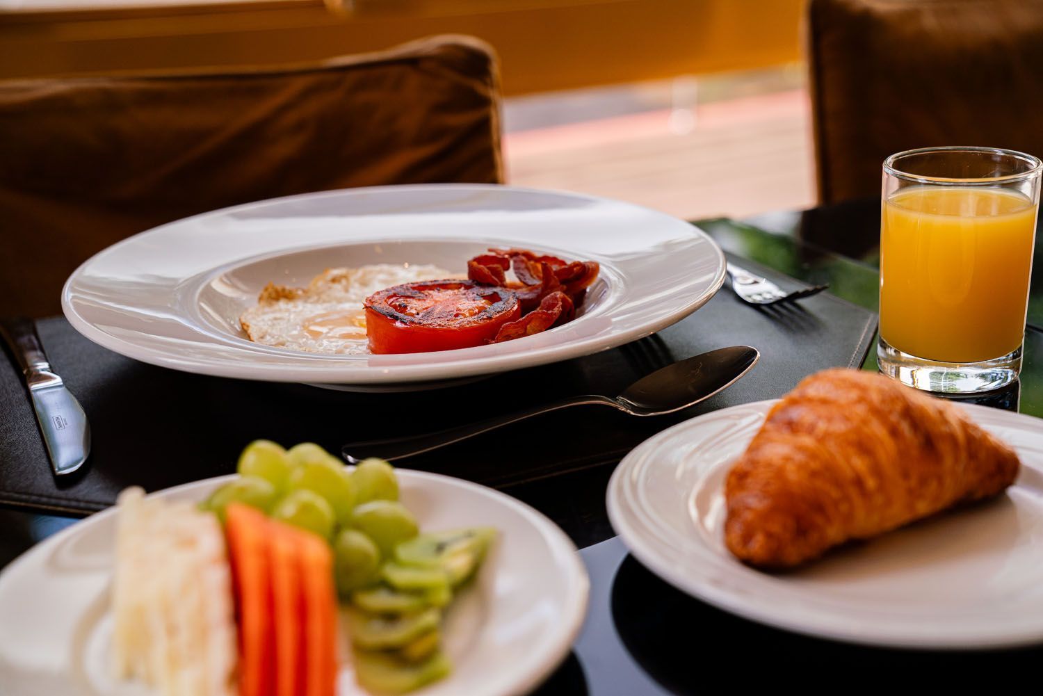 Mesa de café da manhã com prato de comida parcialmente consumido, prato de frutas, croissant e suco de laranja.