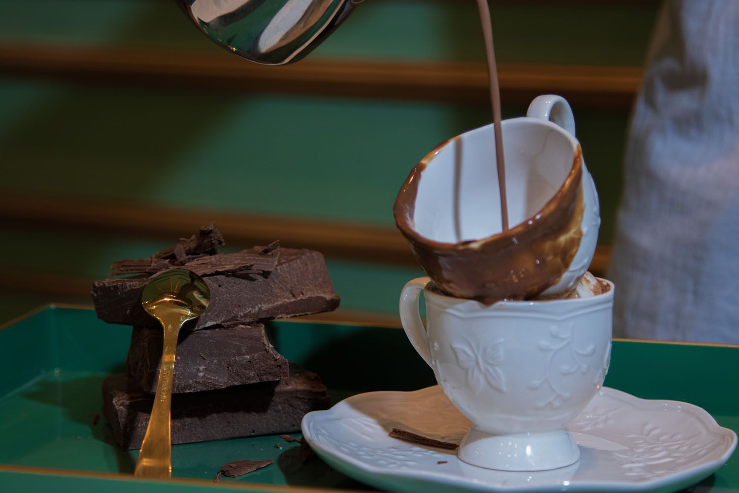Chocolate quente sendo servido em xícaras de chá empilhadas sobre um pires, ao lado de uma colher de ouro e barras de chocolate.