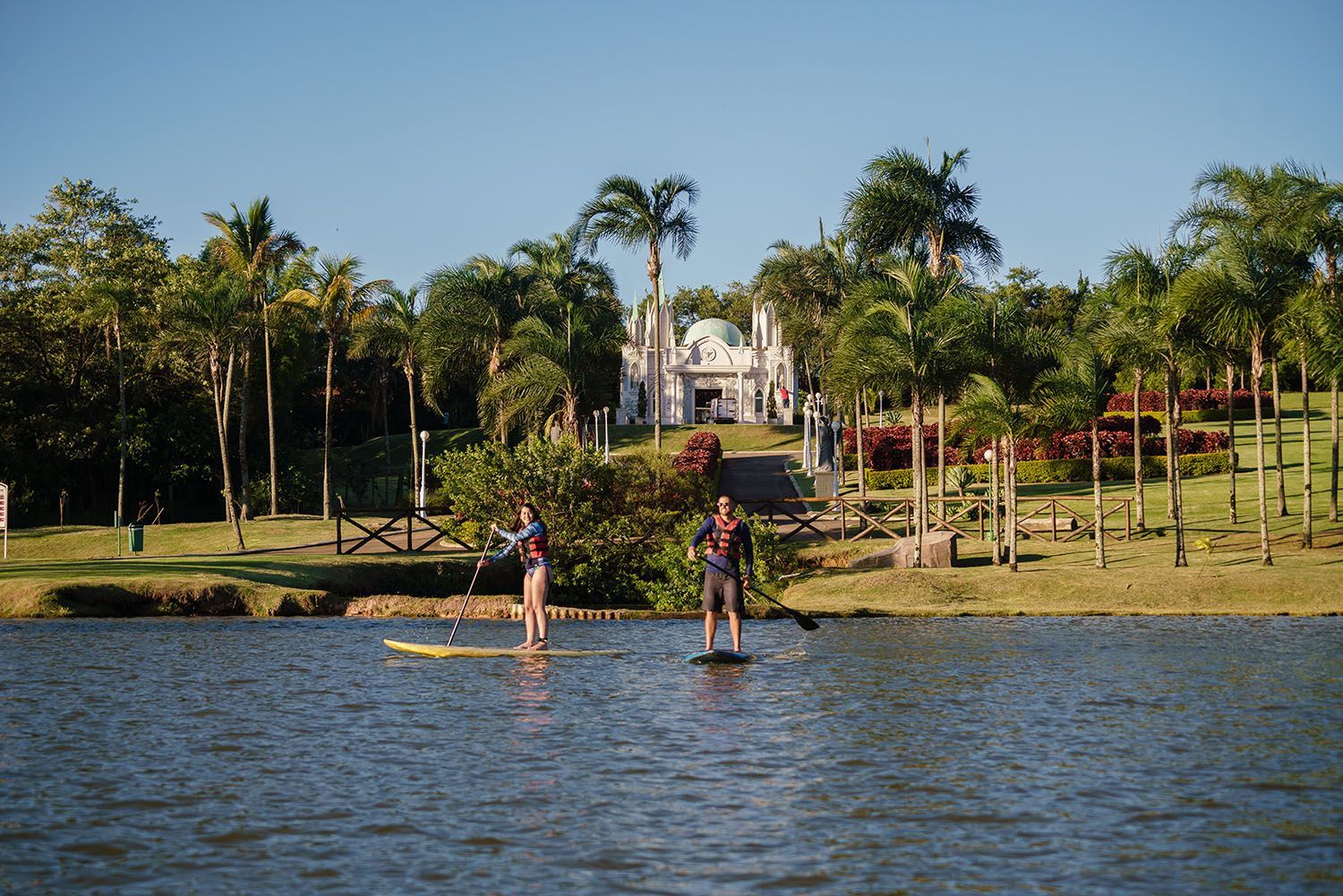 Duas pessoas praticam stand up paddle em um lago, com uma igreja e árvores ao fundo sob um céu ensolarado.