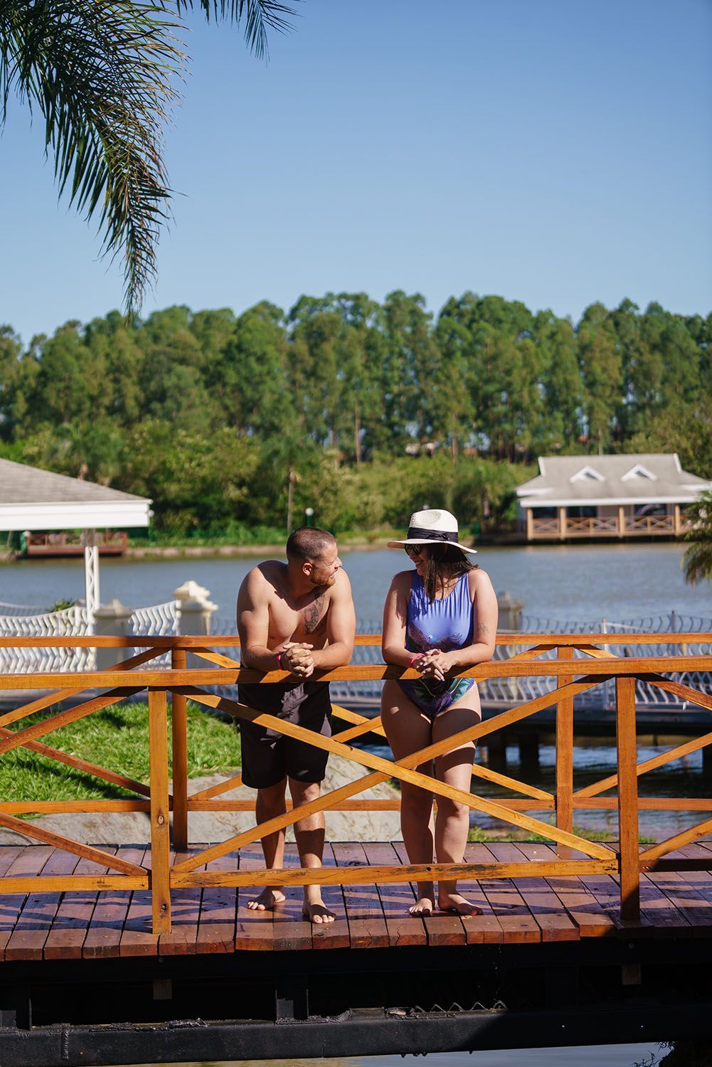 Homem e mulher em uma ponte de madeira sobre um lago. O homem está sem camisa, esfregando os braços. A mulher usa chapéu e traje de banho. Árvores verdes ao fundo.