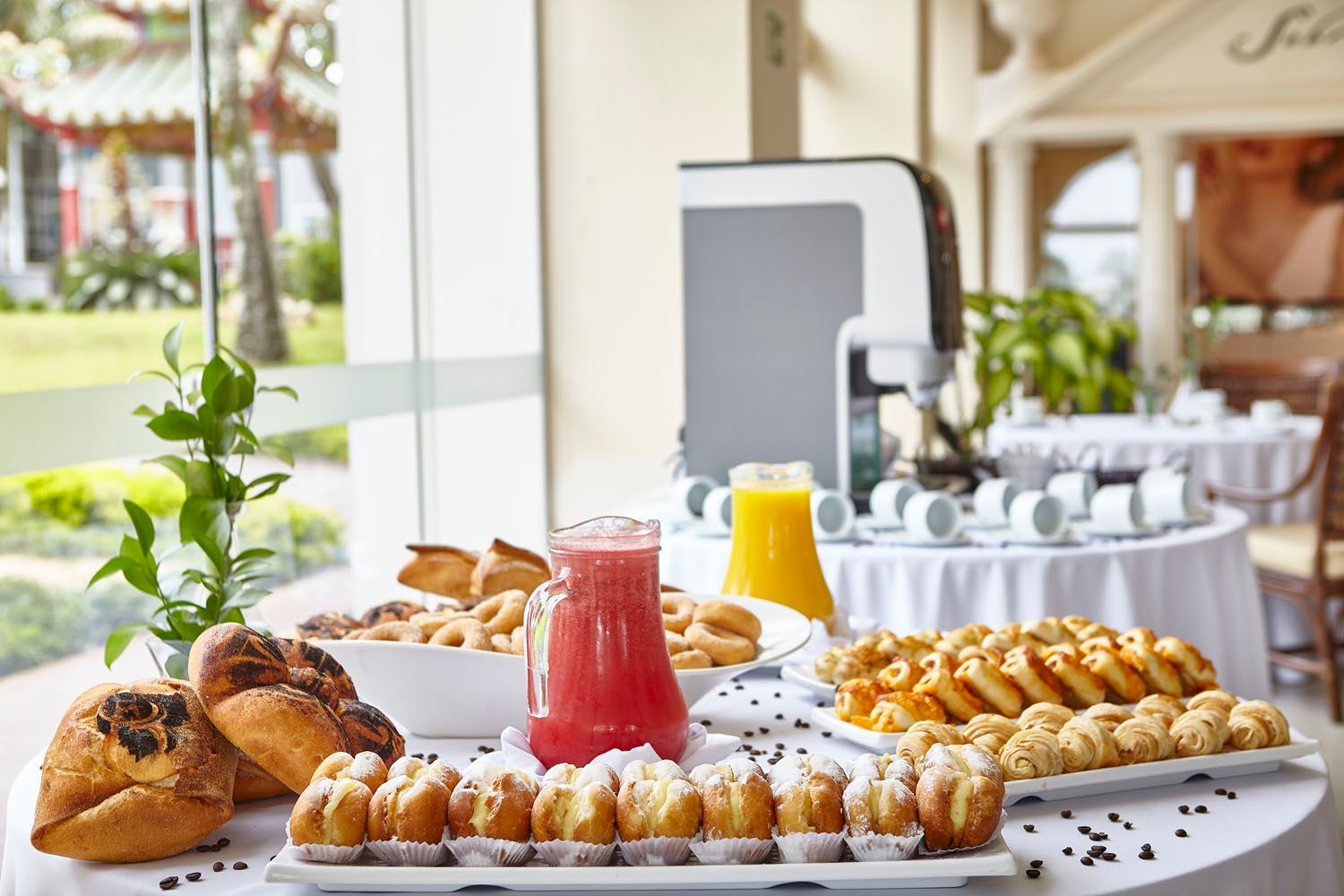 Buffet de café da manhã com doces, sucos e cafeteira. Quarto iluminado com vista para o exterior.