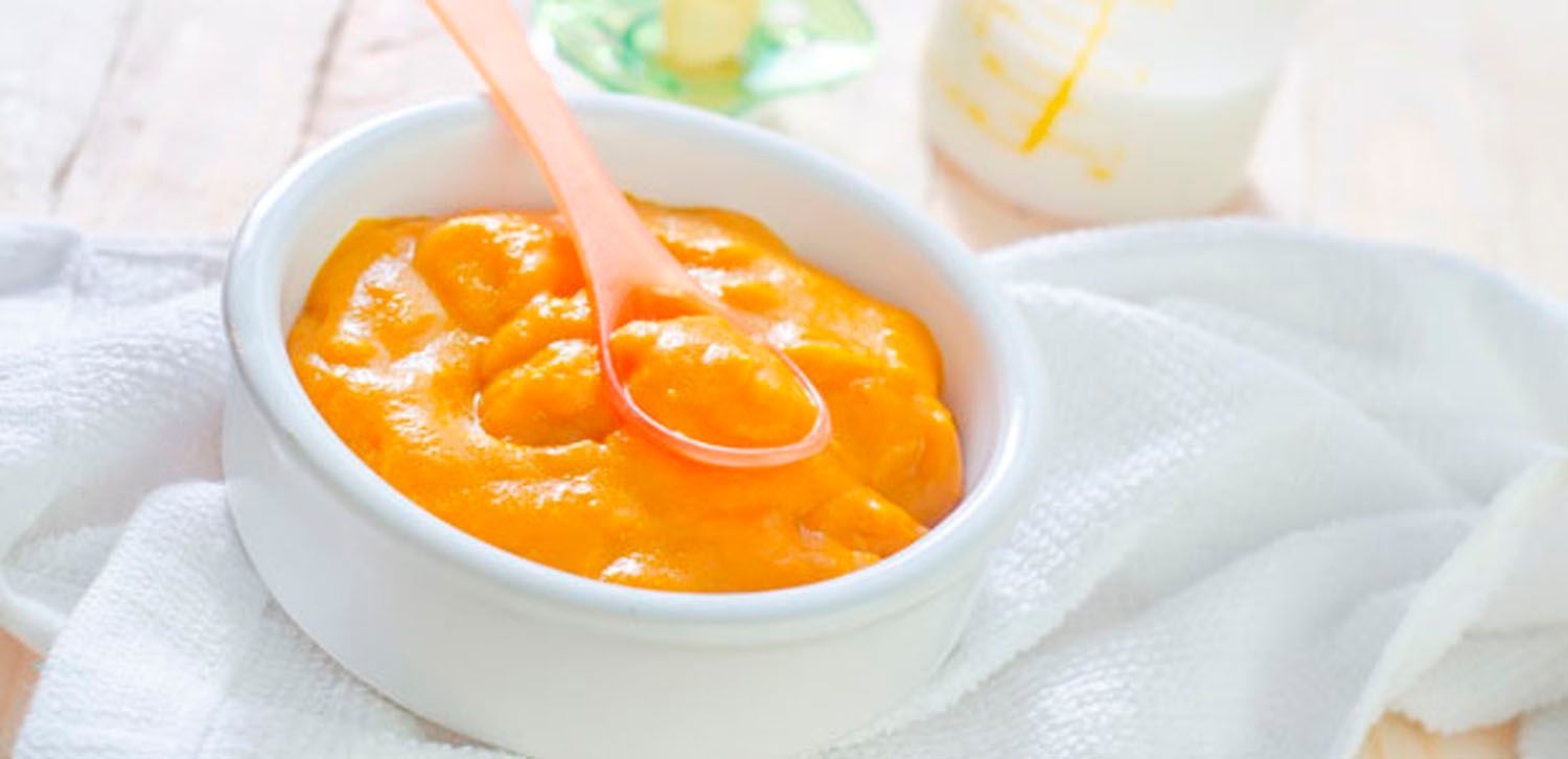 Tigela de papinha laranja com uma colher sobre um pano branco; uma mamadeira ao fundo.