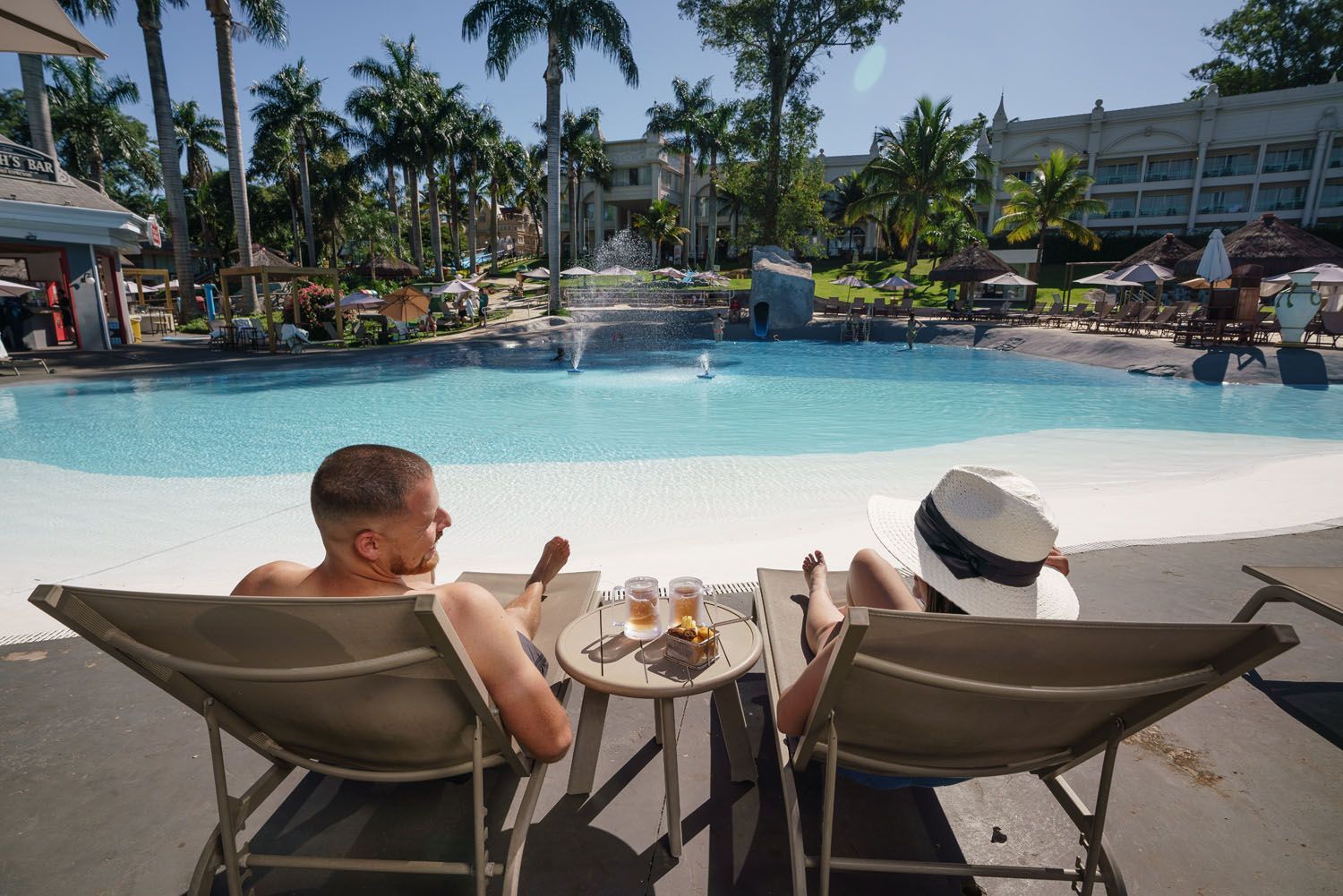 Casal relaxando em espreguiçadeiras à beira da piscina com petiscos e bebidas. Ambiente tropical com palmeiras.