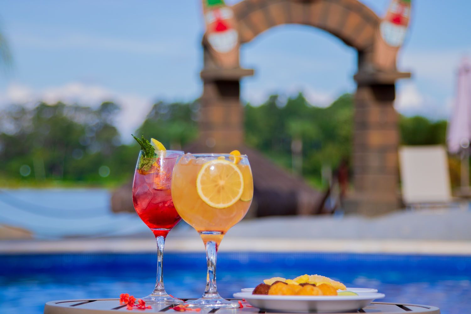 Dois coquetéis com guarnição em uma mesa perto da piscina, com um arco ao fundo.