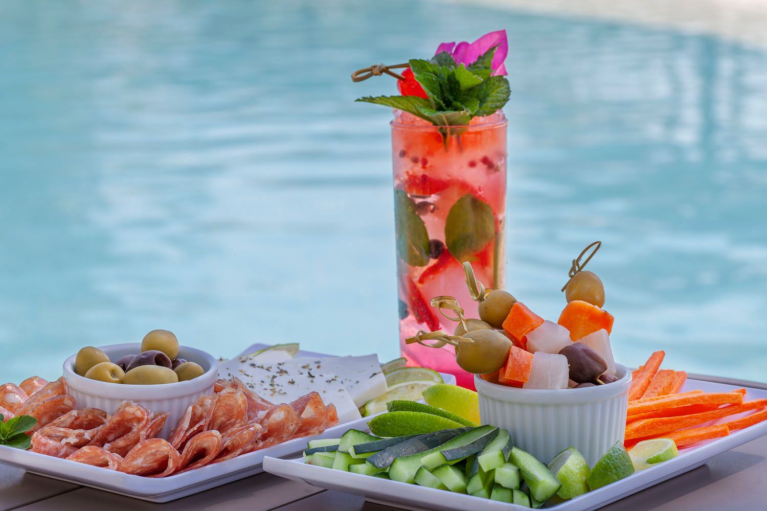 Coquetéis, lanches e aperitivos perto da piscina; frios, azeitonas, espetos, vegetais e bebidas estão em uma bandeja.