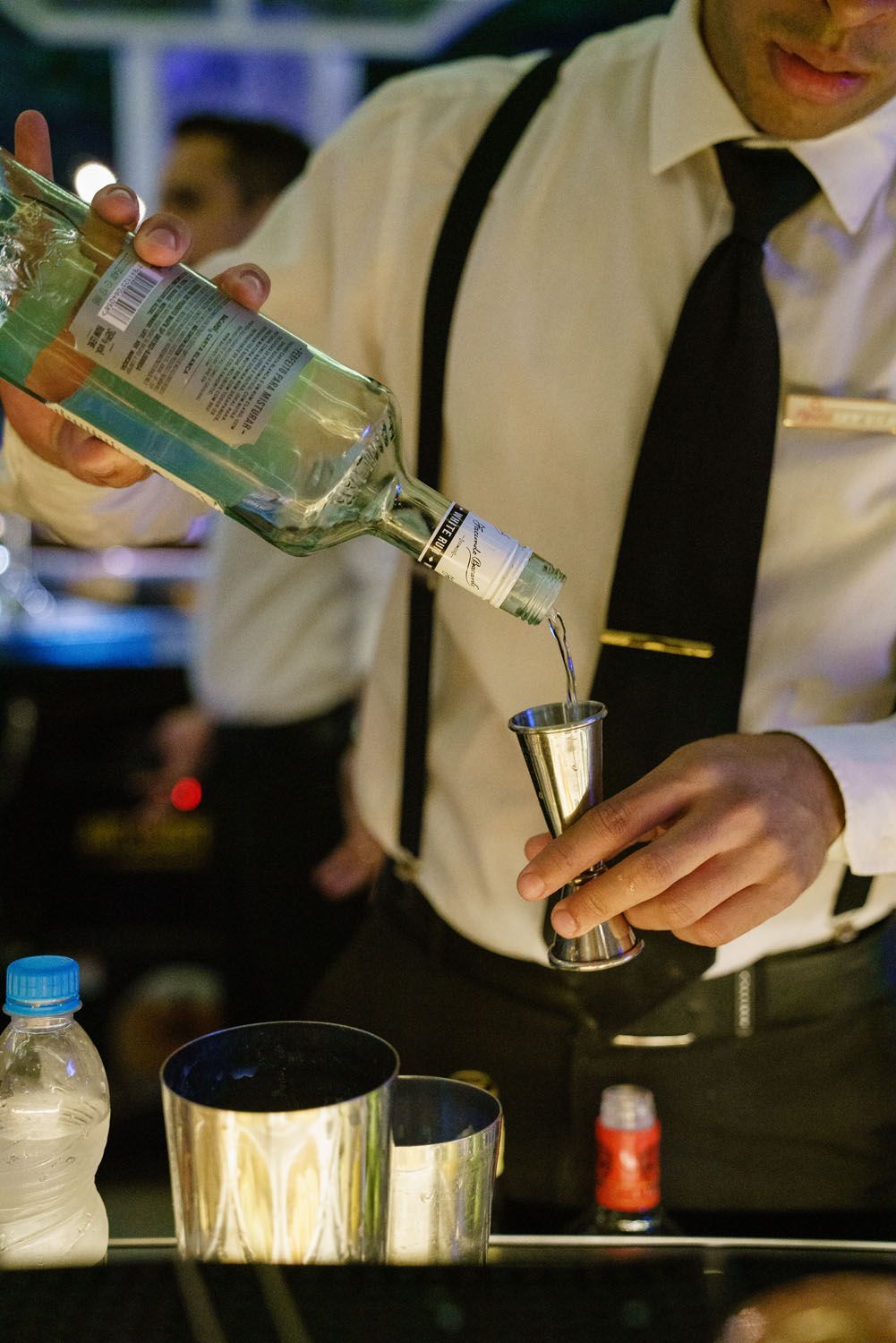 Barman despejando um líquido turquesa de uma garrafa em uma coqueteleira de prata.