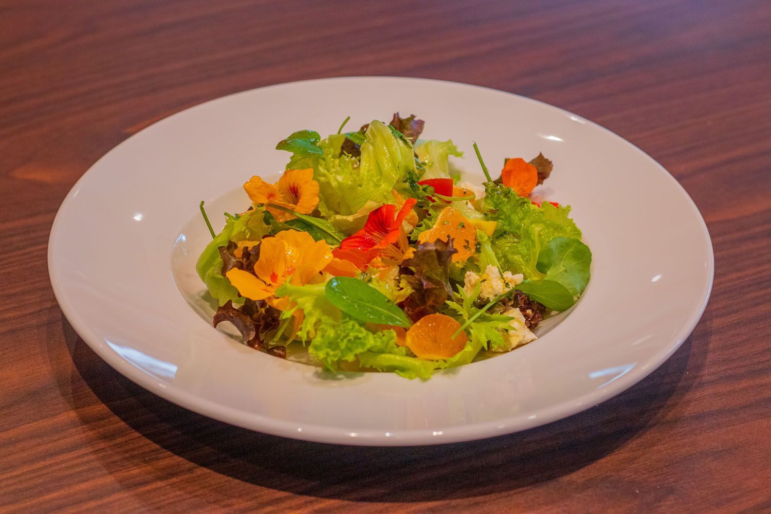 Salada em uma tigela branca sobre uma mesa de madeira; inclui alface, flores, fatias de laranja e pimentões vermelhos.