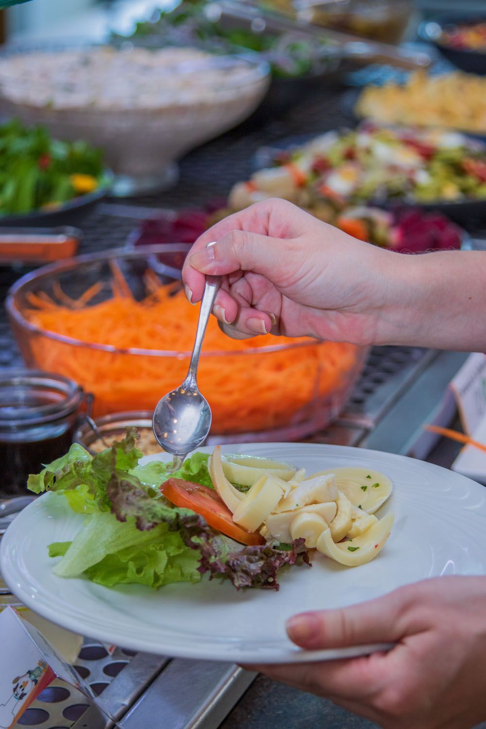 Pessoa adicionando molho a uma salada em um prato em um bufê, com várias opções de comida ao fundo.