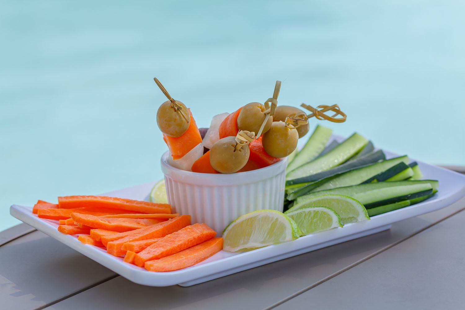 Prato de legumes com cenouras, pepino, limas e espetos de azeitonas, perto de uma piscina.