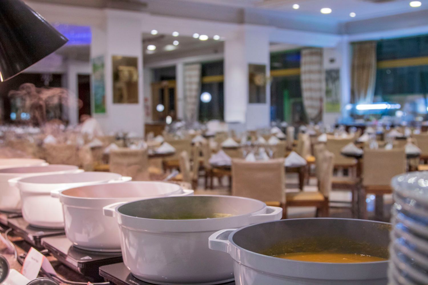 Buffet com tigelas brancas de comida em frente a mesas com toalhas brancas.