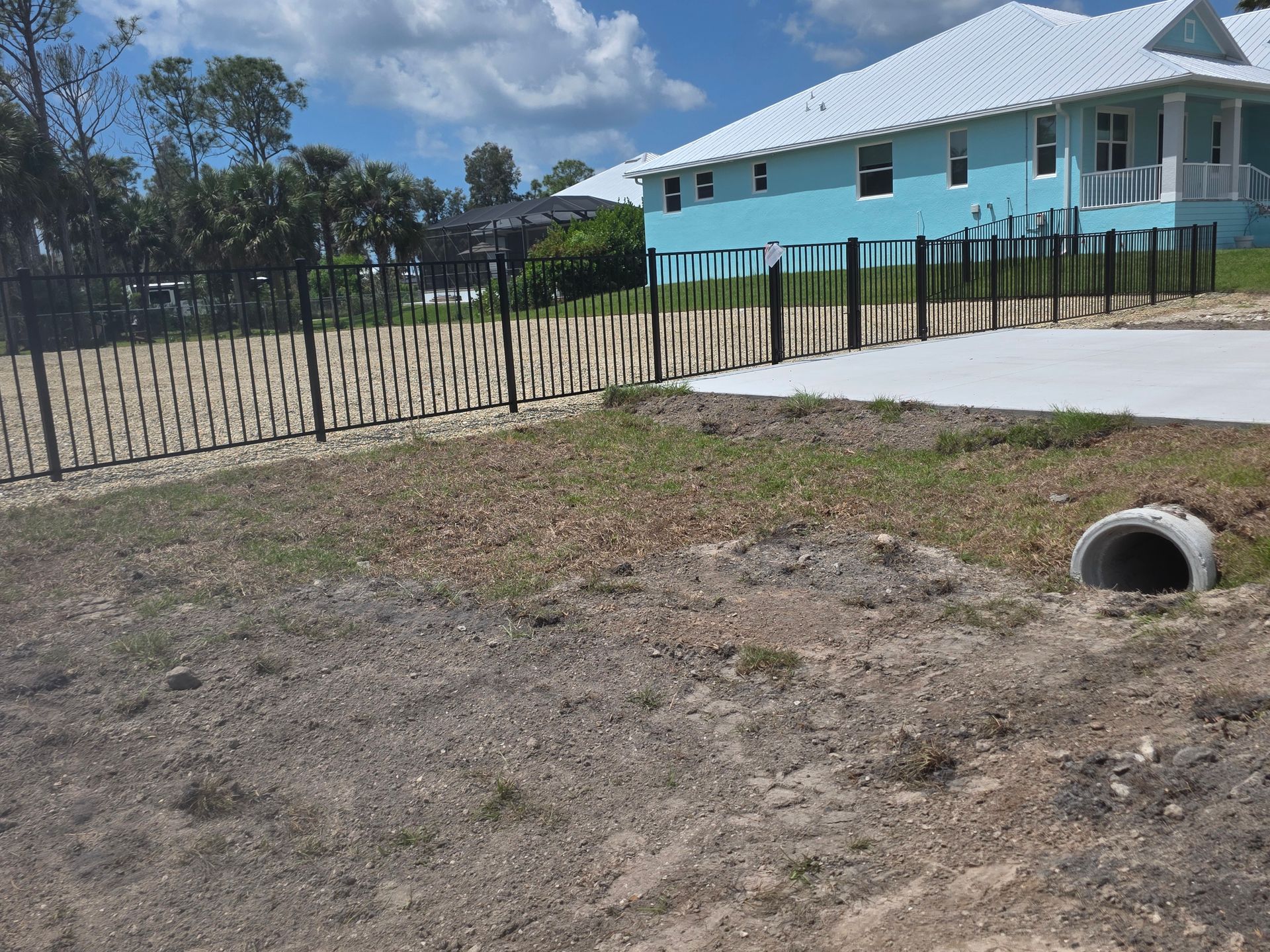 Vacant sandy lot beside a turquoise house with a wooden fence and drainage pipe under a cloudy sky