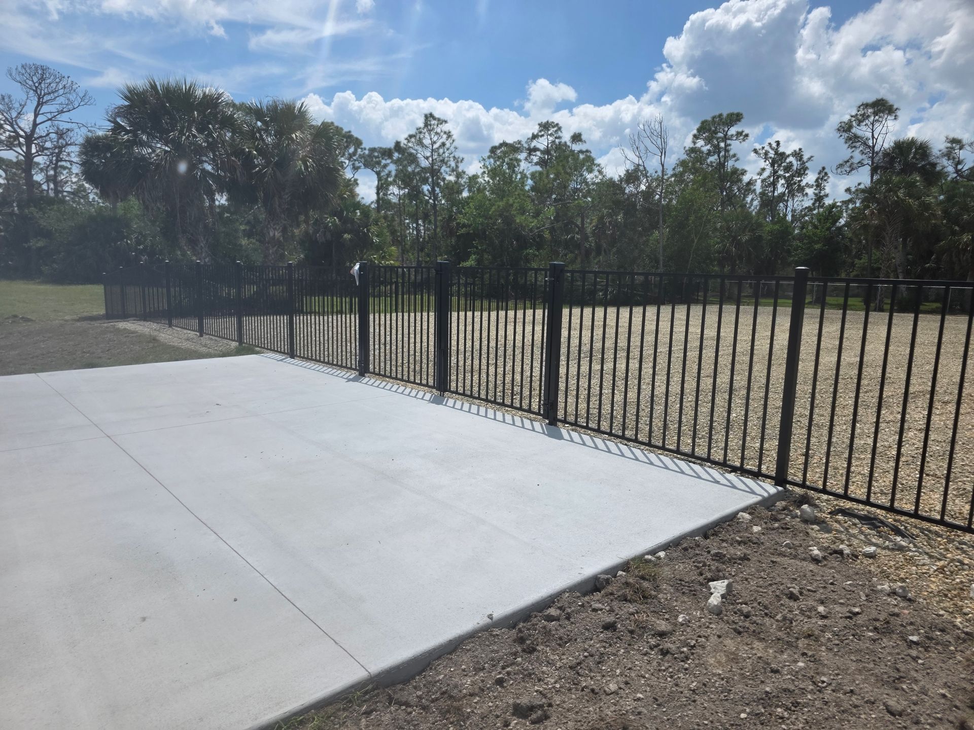 Concrete patio beside black metal fence and dry grassy field under a partly cloudy sky