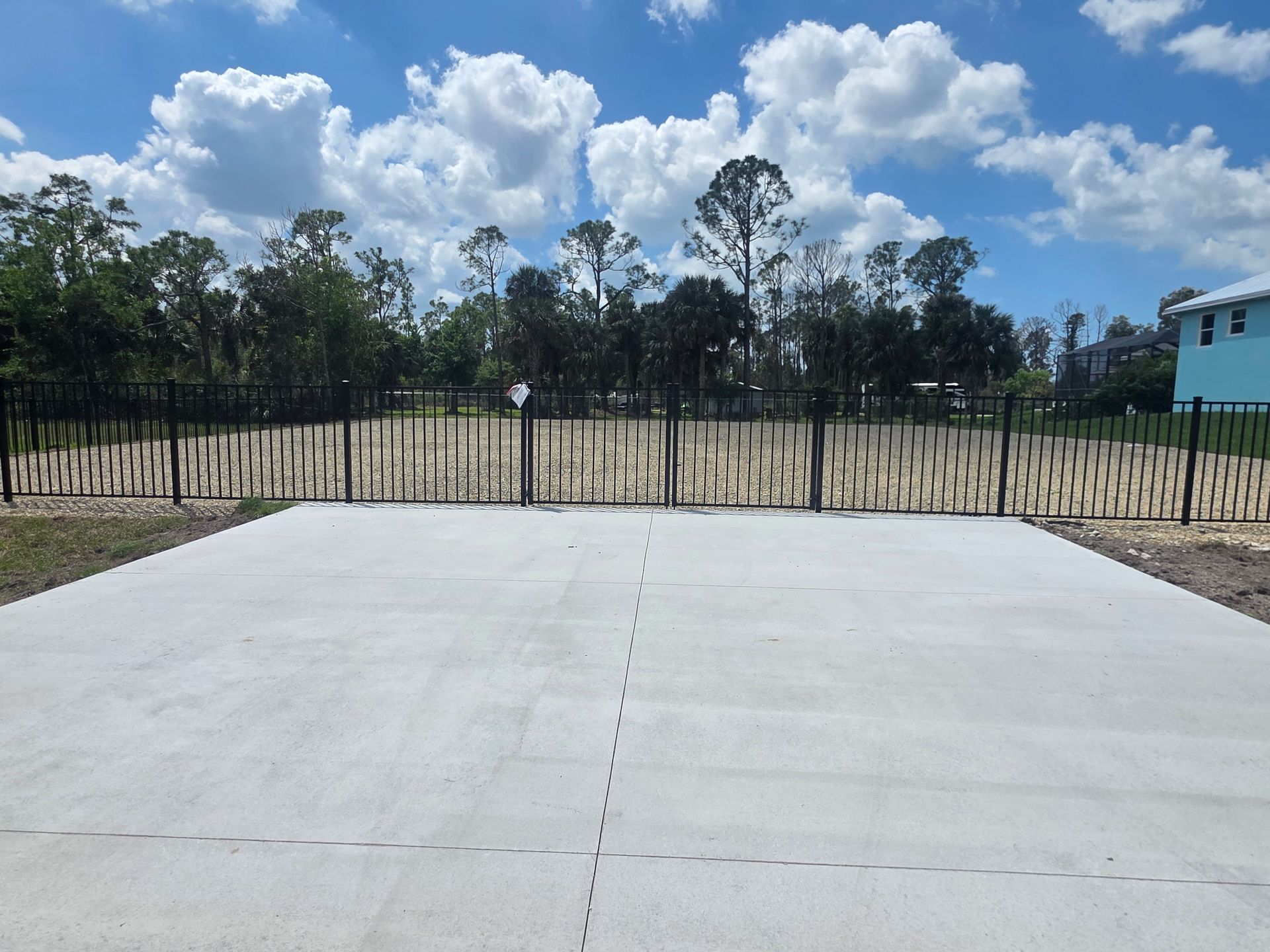 Concrete driveway leads to a fenced yard with trees and a blue building under a partly cloudy sky.