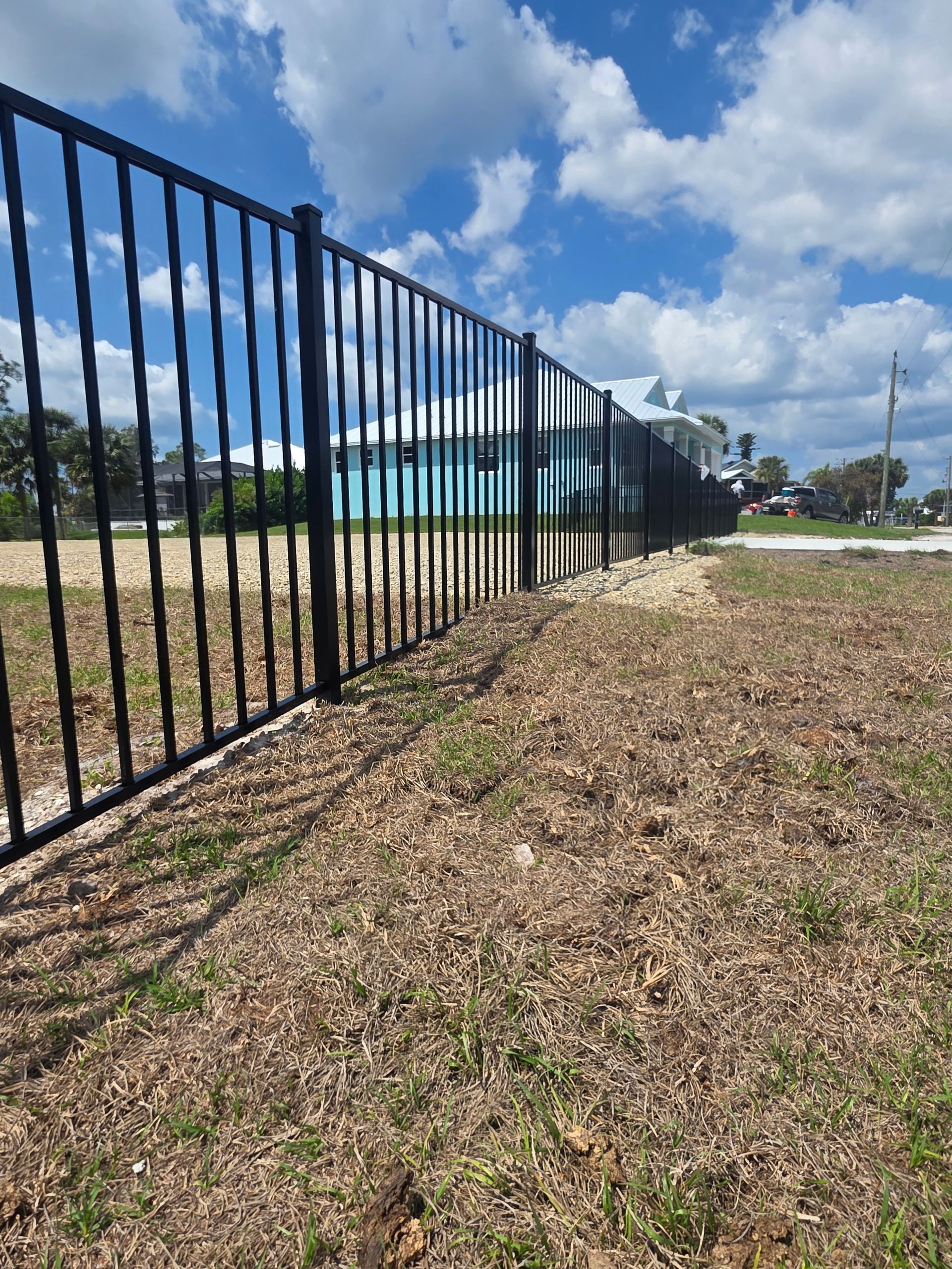 Black metal fence beside a grassy dirt path under a bright blue sky with clouds