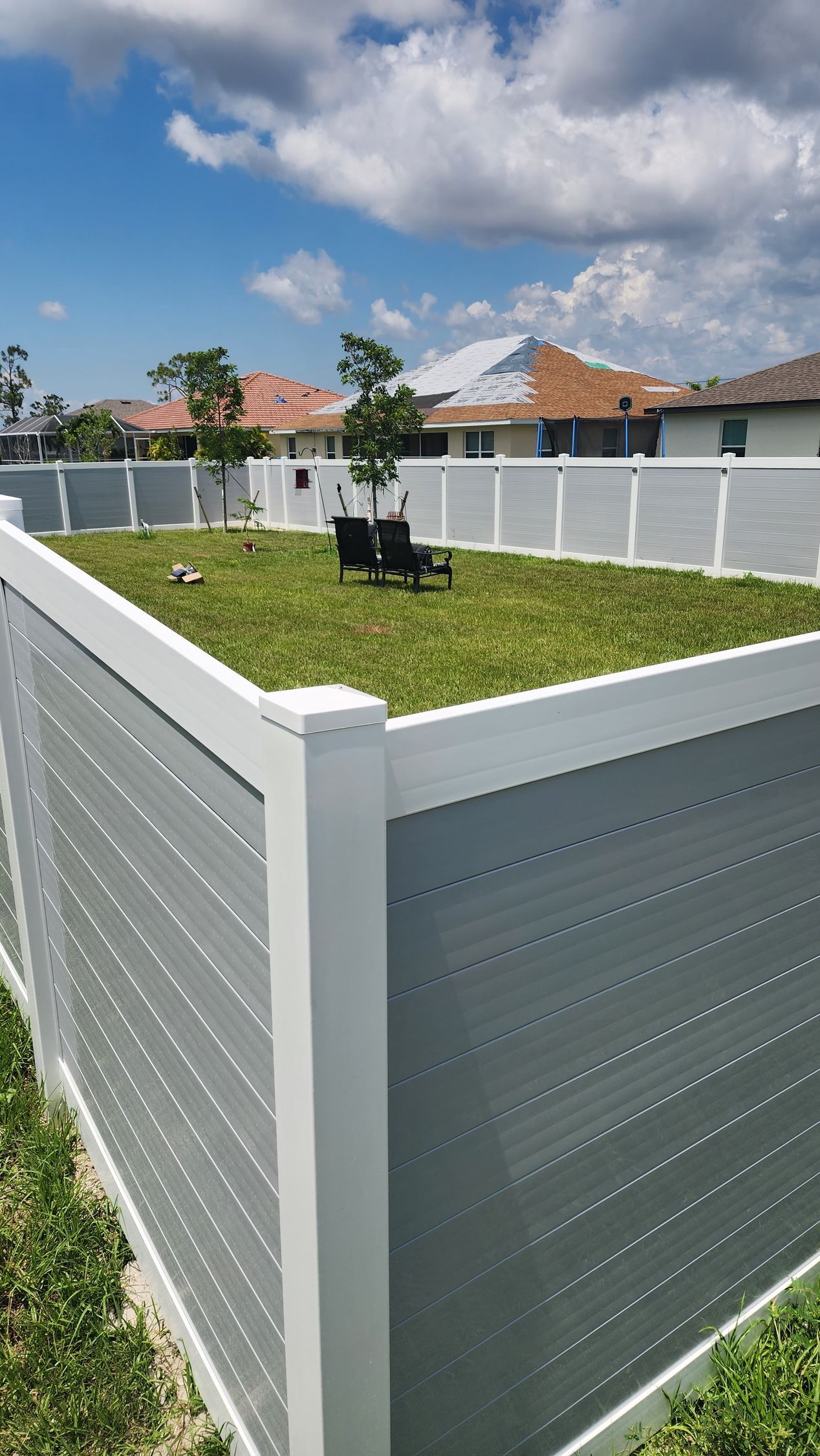 Gray and white fenced backyard with green grass, a few trees, and houses in the background under a blue sky.