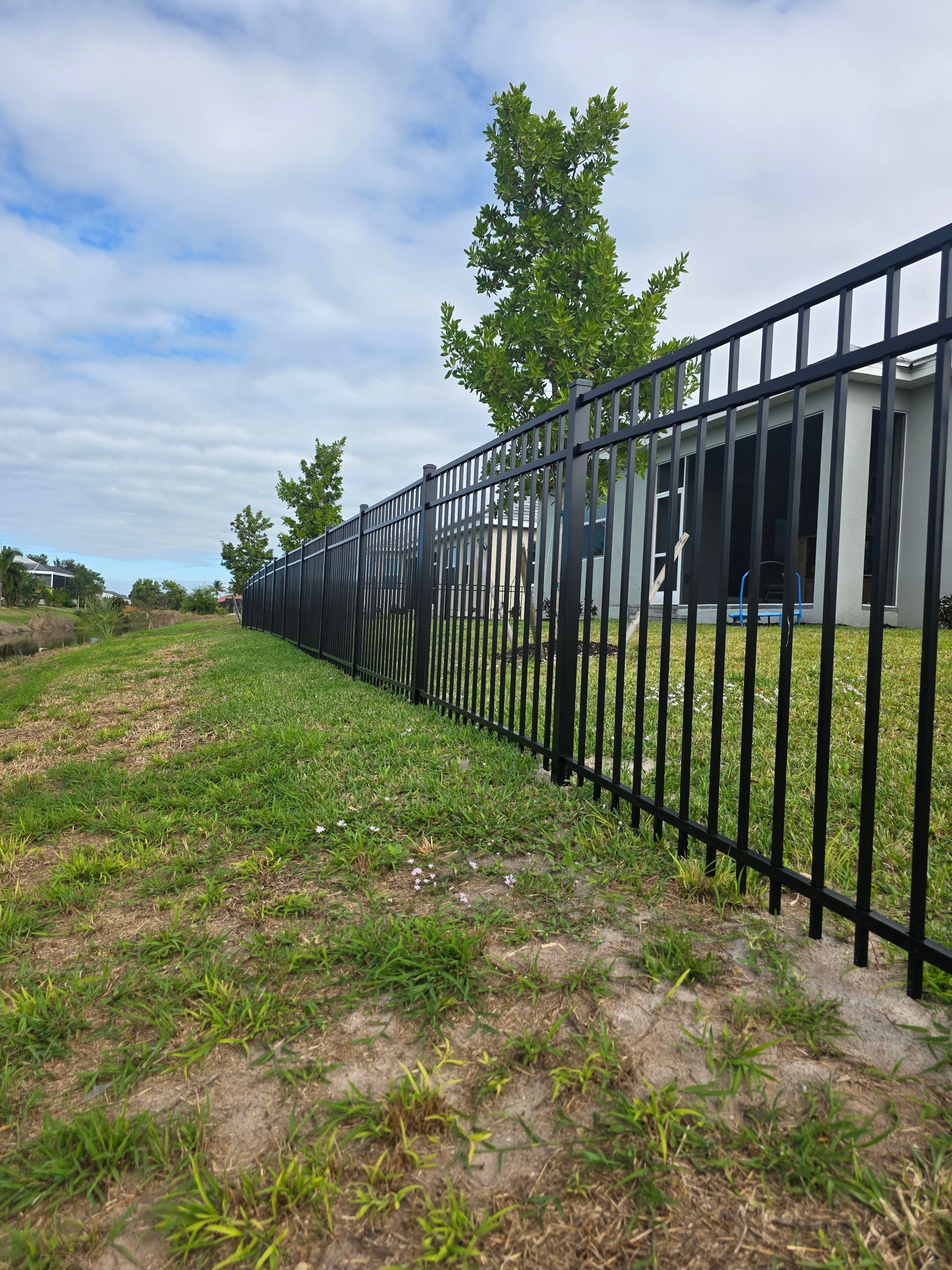 Black metal fence along a grassy area, with a house and trees in the background under a cloudy sky.