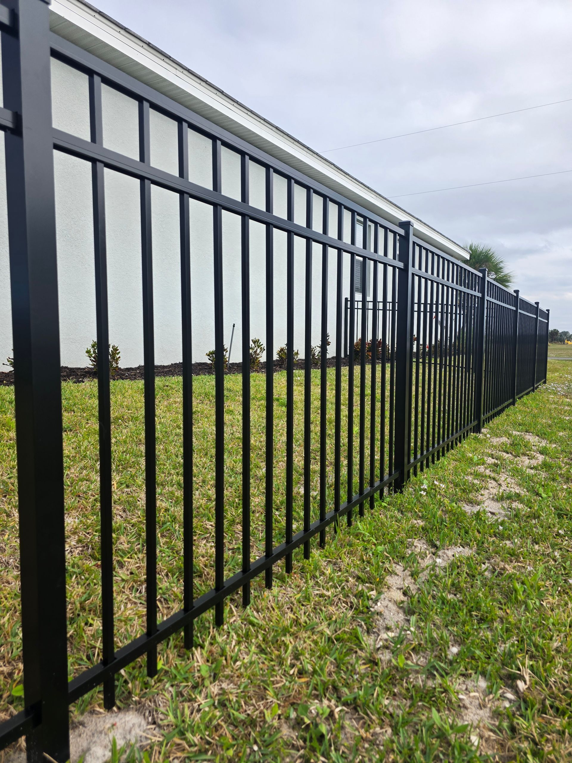 Black metal fence in a grassy yard, beside a white house, under a cloudy sky.