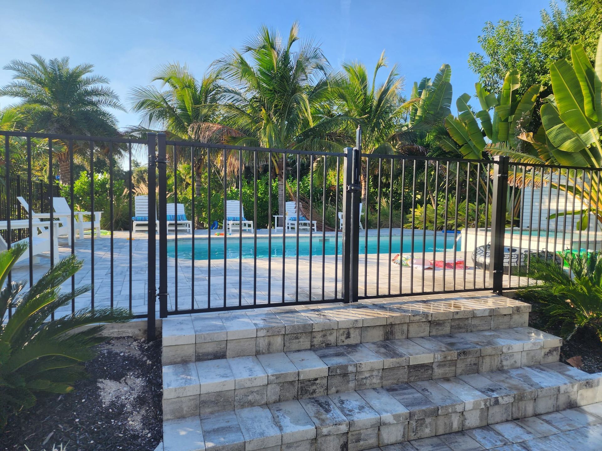Black metal fence around a pool, steps leading up to it, palm trees in background.