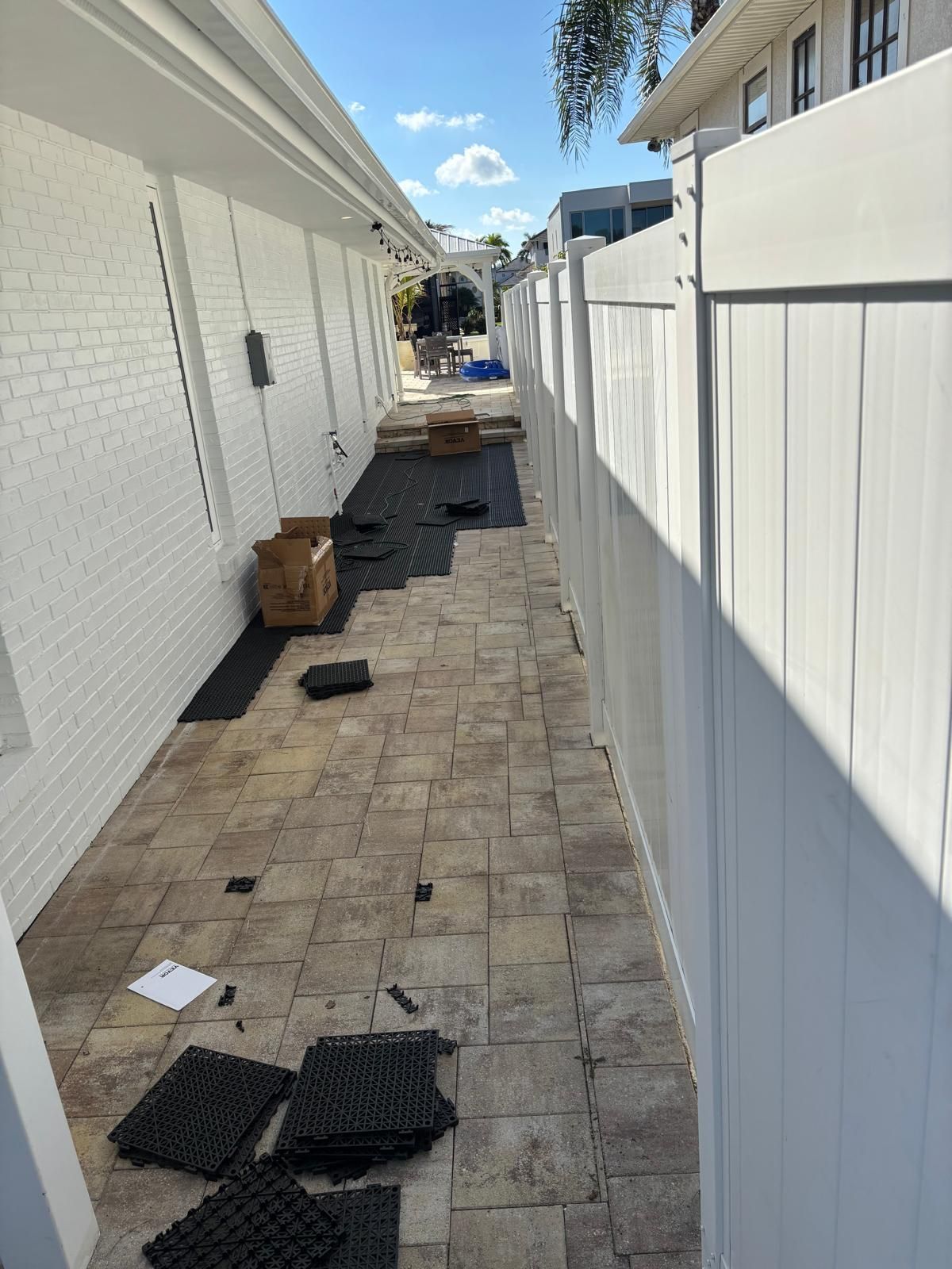 A narrow patio under construction, with pavers and black tiles between a white fence and a white brick wall.