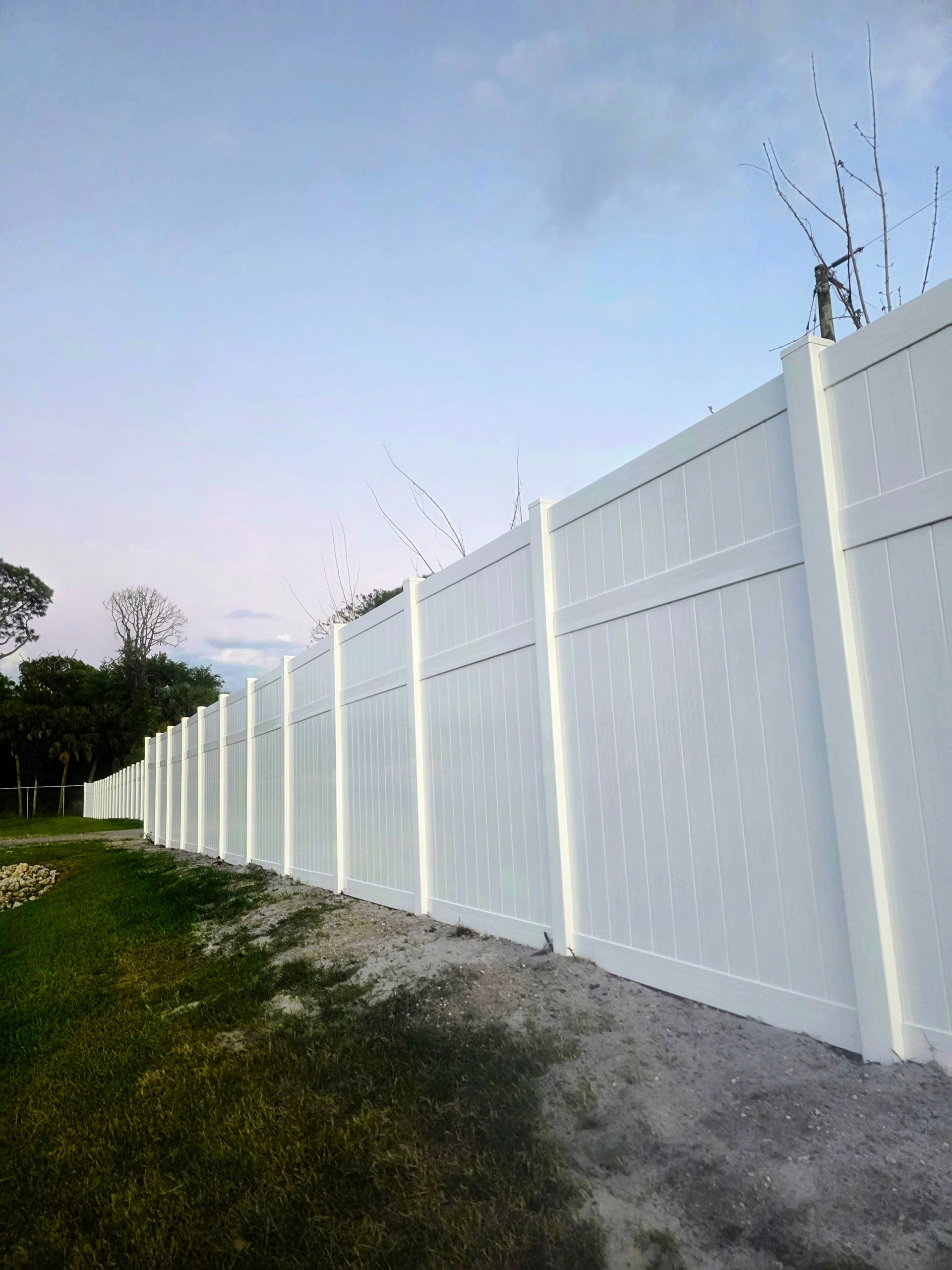 White vinyl privacy fence along a grassy roadside under a pale blue sky.