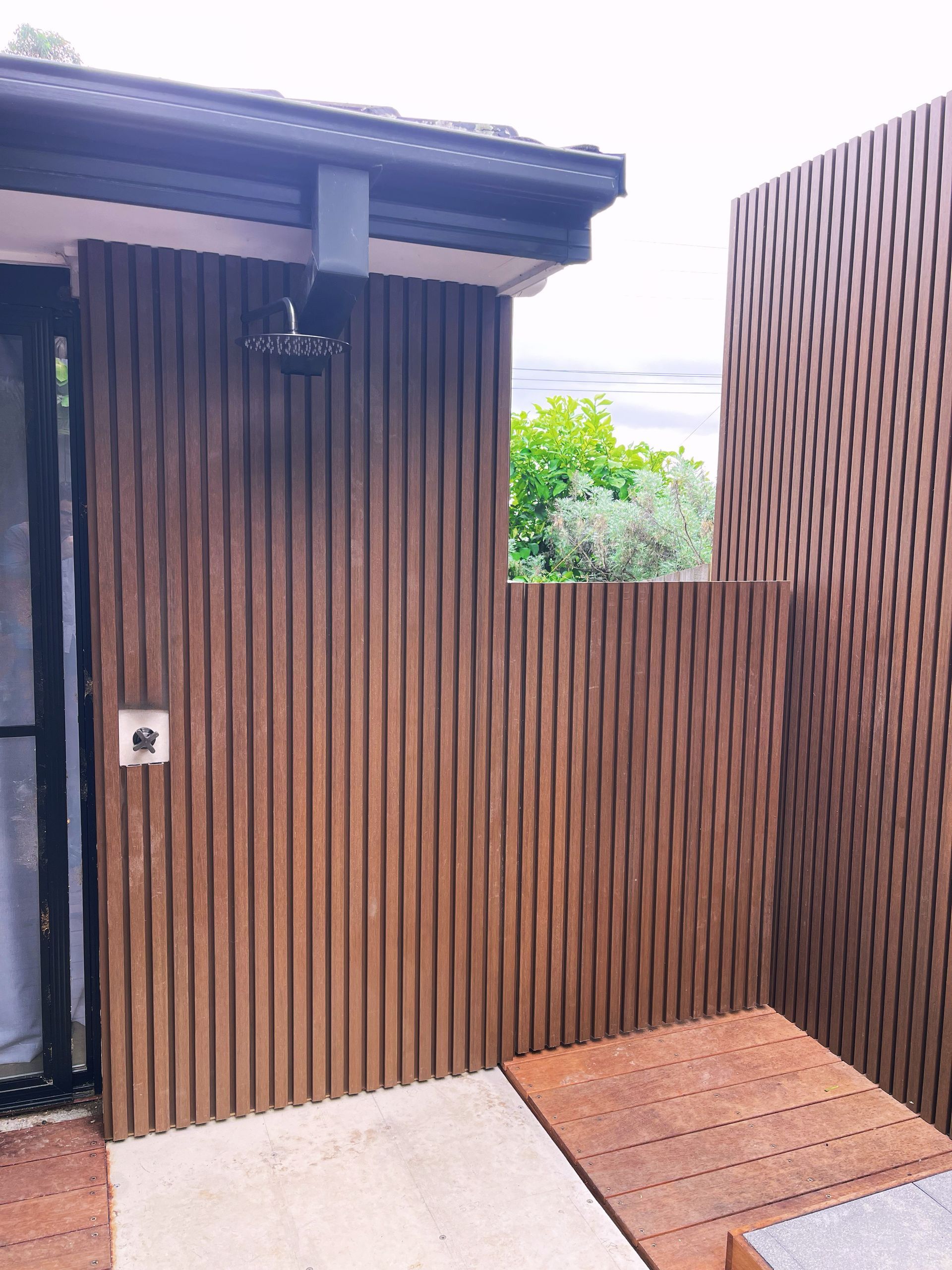 Wooden entrance gate and wall with concrete walkway and small deck, greenery visible beyond the opening