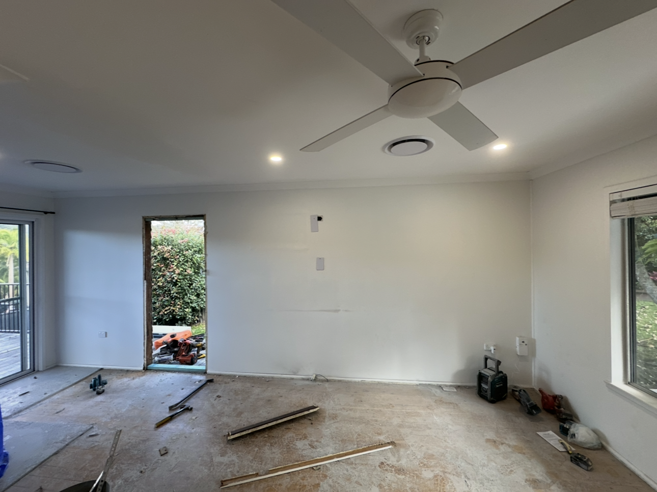 Empty living room with white walls, ceiling fan, carpet debris, and sliding doors to a patio.