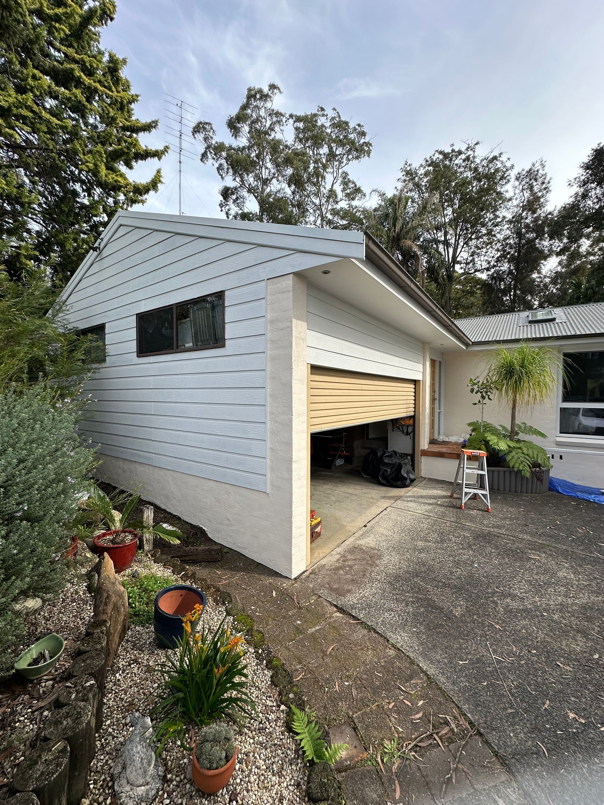 White house garage with open door, gravel driveway, and a parked vehicle beside trees and garden plants