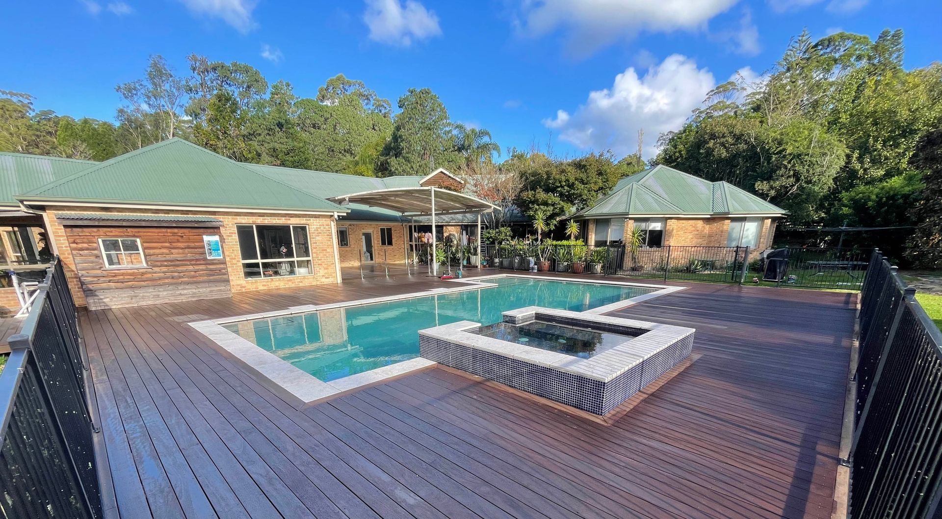 Modern backyard deck with a swimming pool, hot tub, and wooden cabanas under a blue sky