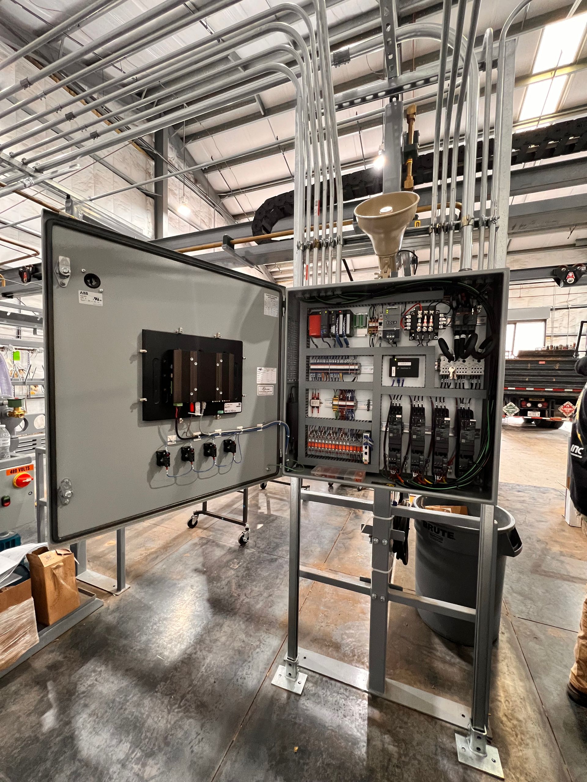 Open industrial electrical control panel with exposed wiring inside a large manufacturing warehouse.
