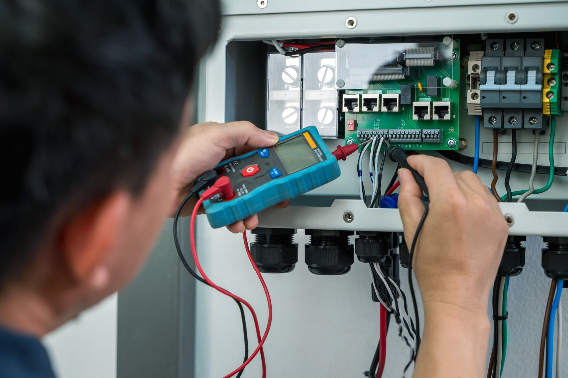 Electrician's hands using a digital multimeter tool to test wiring voltage on a circuit board panel.