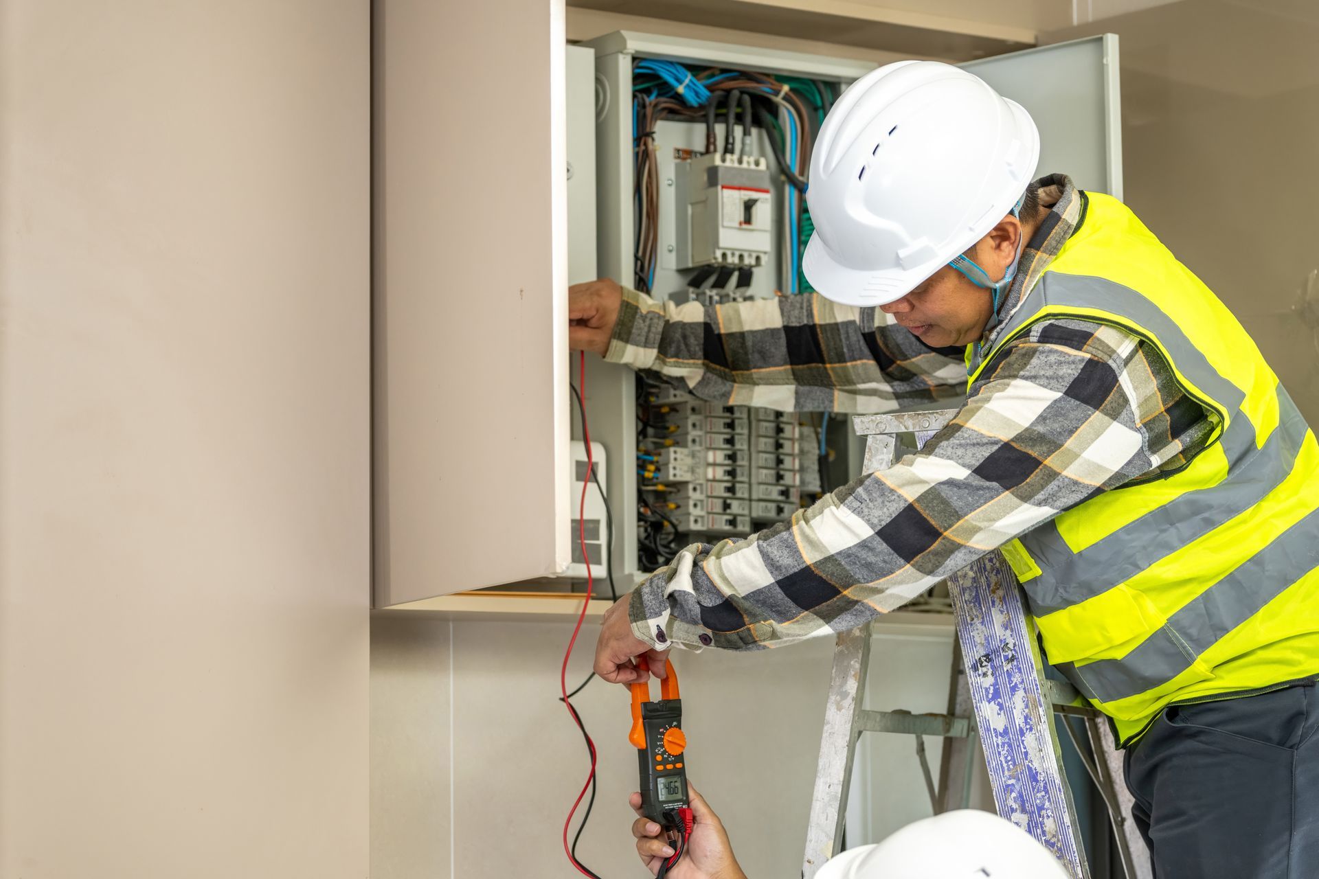 Electrician wearing a hard hat and safety vest testing a building breaker panel with a multimeter.