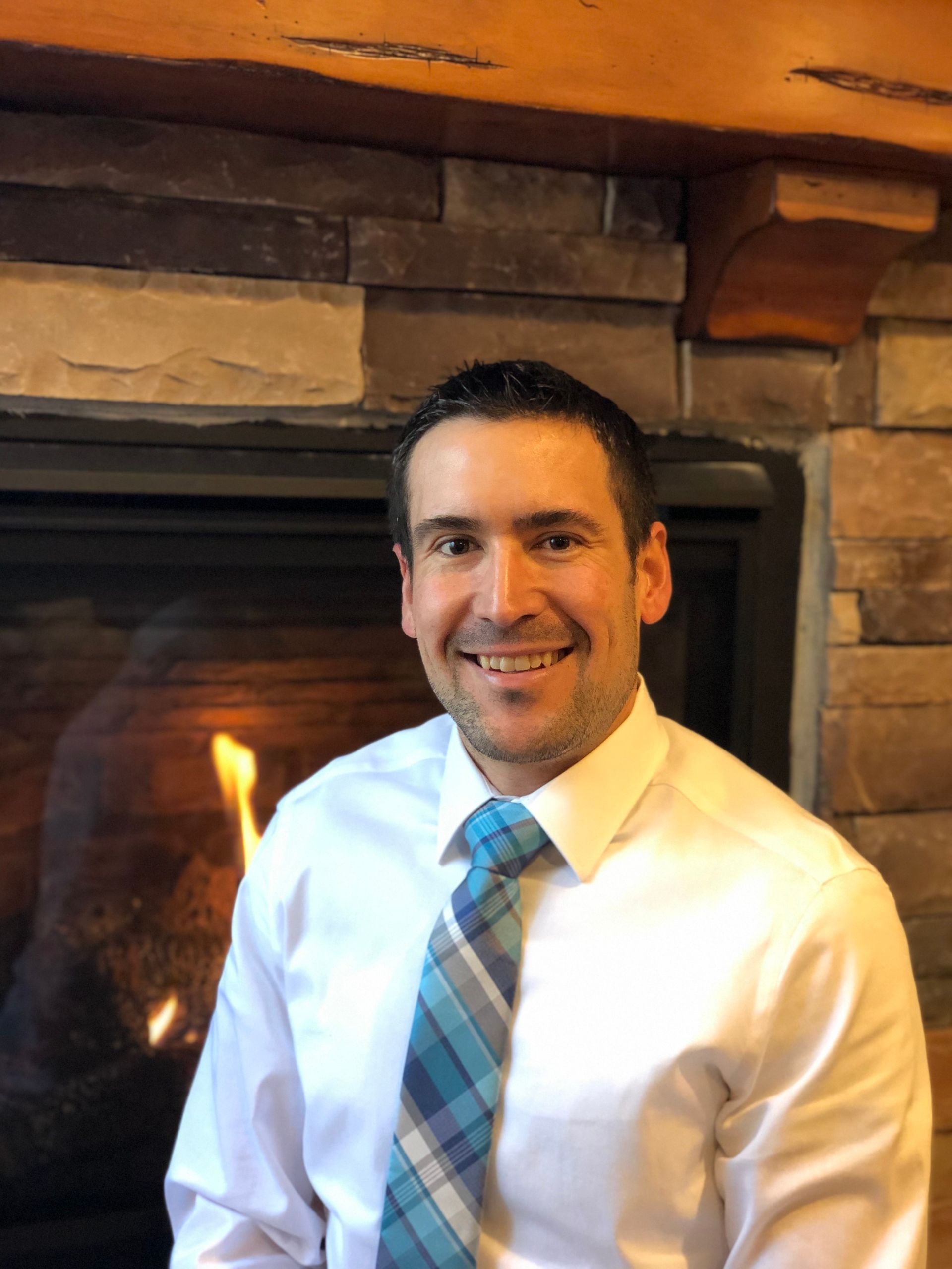 A man in a white shirt and tie is smiling in front of a fireplace.