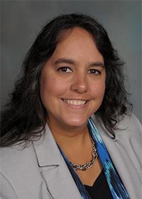 A woman wearing a gray jacket and a blue scarf is smiling for the camera.