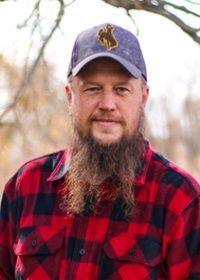 A man with a beard is wearing a plaid shirt and a hat.