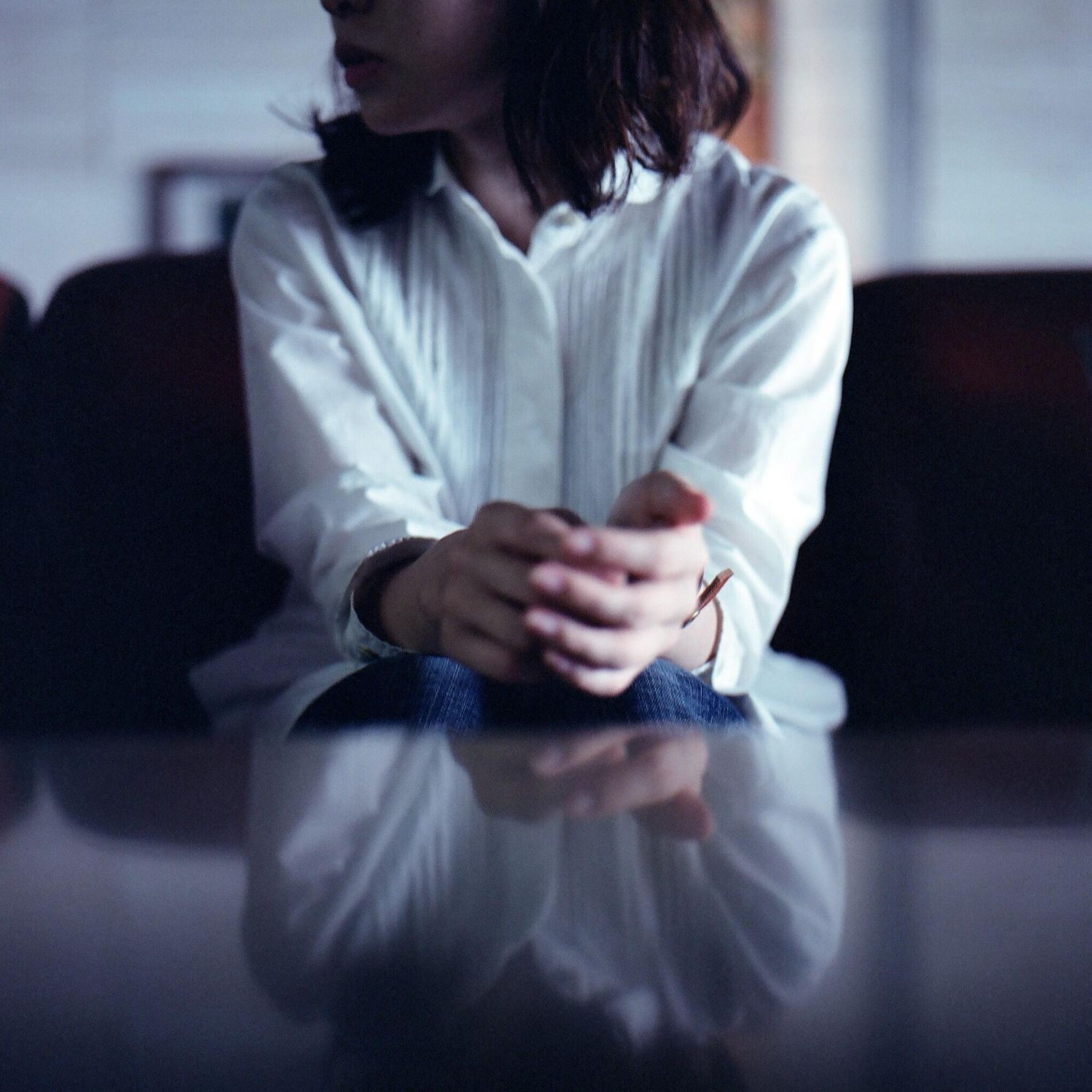 Person in a white shirt sits with clasped hands at a table, reflected on the glossy surface.