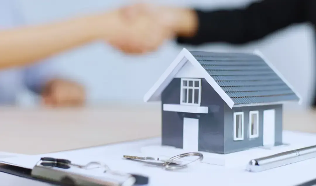A couple of people shaking hands over a model house on a clipboard.
