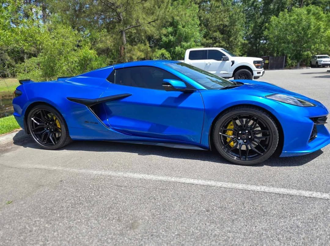 Blue sports car parked on asphalt, black wheels, yellow brake calipers, white truck in the background.