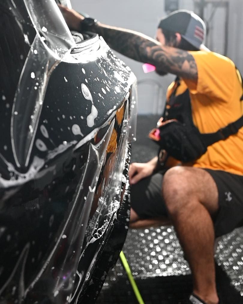 Person washing a black car with soap suds in a car wash, wearing an orange shirt and black cap