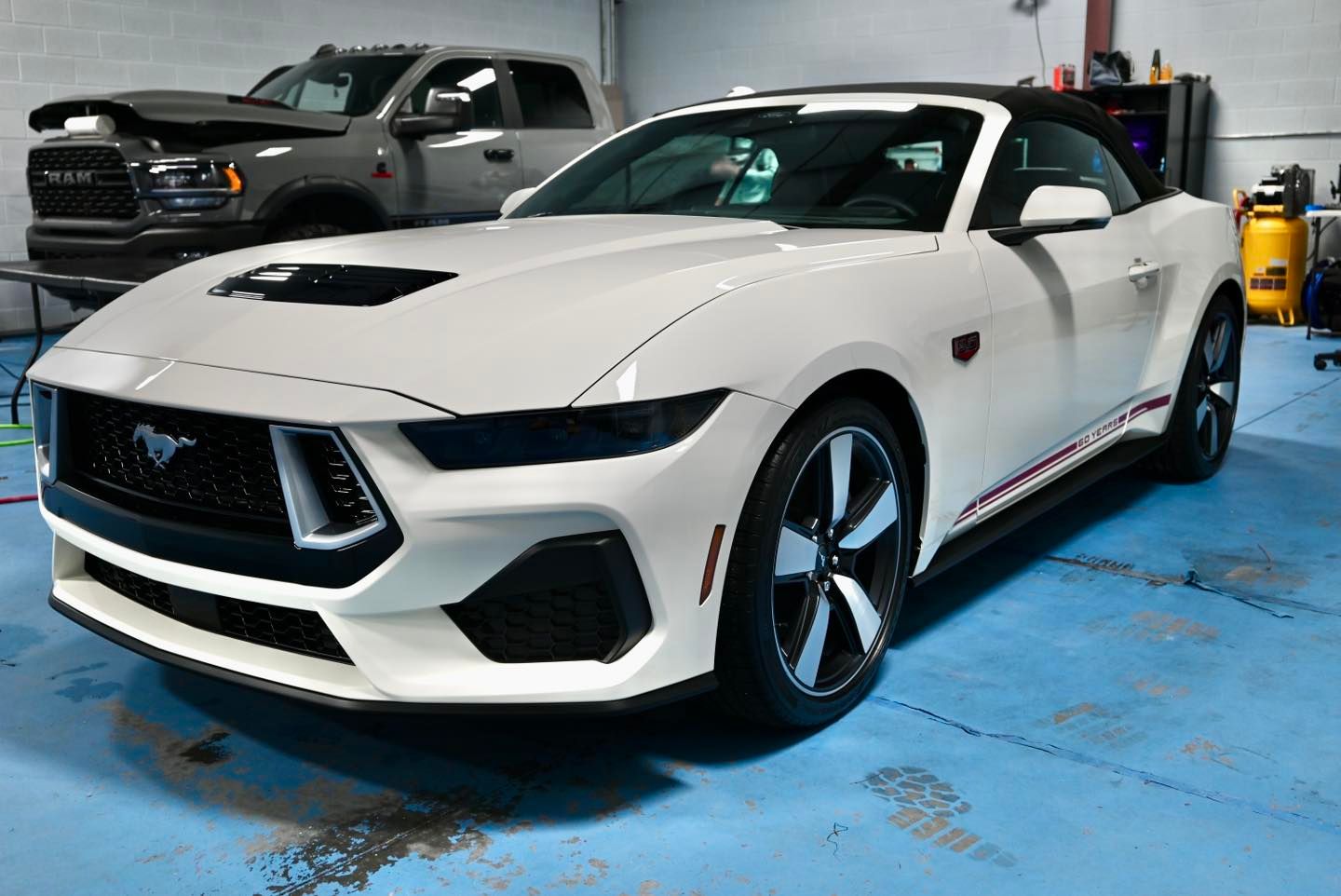White Ford Mustang convertible with black accents in a garage, with a Ram truck in the background.