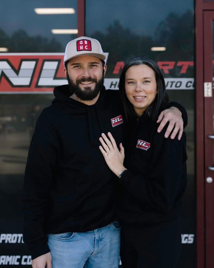 Two people in black hoodies pose smiling outside a storefront with a red-and-black sign.