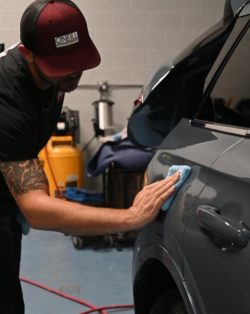Person polishing a gray car with a cloth in a garage, wearing a red cap and black shirt.